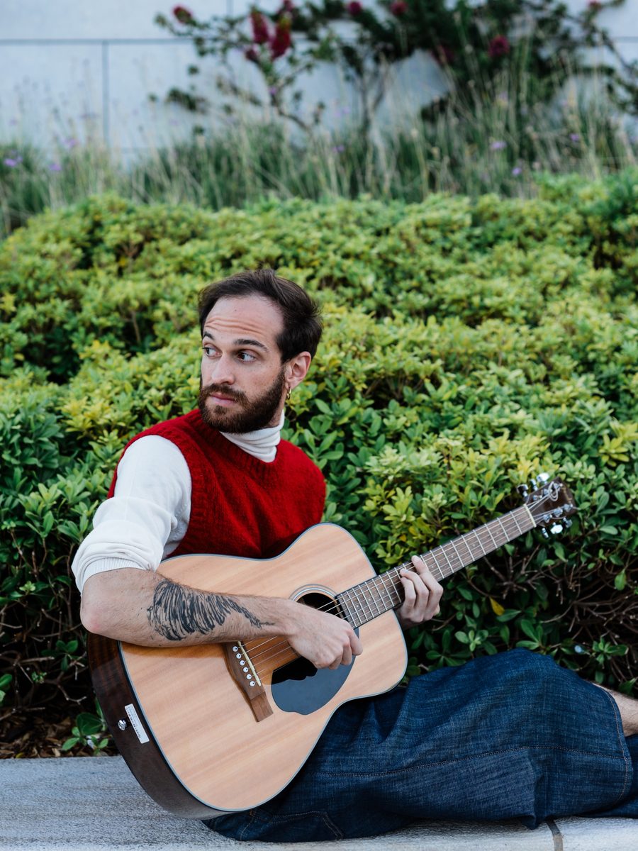 Mochita sitting outdoors with his acoustic guitar, in front of a green hedge